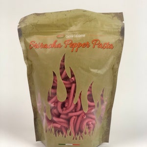 Puede incluir: Una bolsa de papel marrón con una ventana transparente que muestra pasta roja en su interior. La bolsa tiene una etiqueta que dice "Cucina Calabrese Sriracha Pepper Pasta".