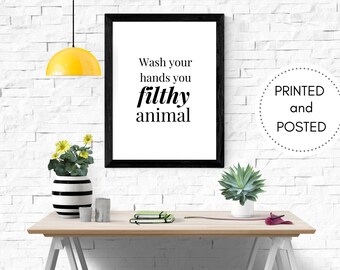May include: A framed print with the text "Wash your hands you filthy animal" in black font. The print is displayed on a wooden table with a yellow lamp, a vase with flowers, and other decorative items.