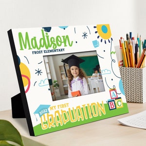 May include: A white picture frame with a colourful design featuring a child in a graduation cap and gown. The frame says "Madison Frost Elementary" and "My First Graduation 2022".