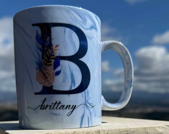 May include: A white ceramic mug with a blue marble design. The mug features a large black letter "B" with floral accents and the name "brittany" written in cursive below the letter.