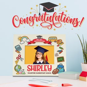 May include: A personalised graduation photo frame with a yellow background and colourful school-themed illustrations. The frame has a space for a photo and includes the text "2022 My 1st Graduation", "Shirley", and "Valentine Elementary School".
