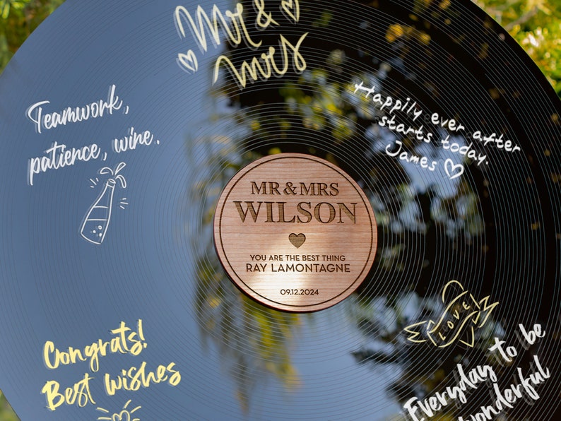 May include: A black vinyl record with gold lettering that says "Mr & Mrs" and "Happily ever after starts today. James." A wooden circle with the text "MR & MRS WILSON" and "YOU ARE THE BEST THING RAY LAMONTAGNE 09.12.2024" is placed on the record.