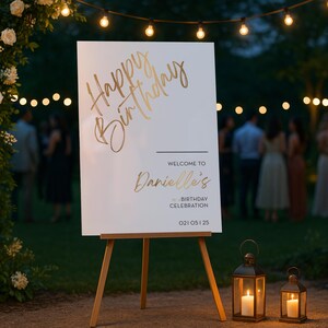 以下が含まれることがあります： 「Happy Birthday」と「Welcome to Danielle's Birthday Celebration」と金色の文字で書かれた、木製のイーゼルに立てられた白い看板。2つのライトアップされたランタンが看板の両側にあり、背景には花のアーチとストリングライトがあります。