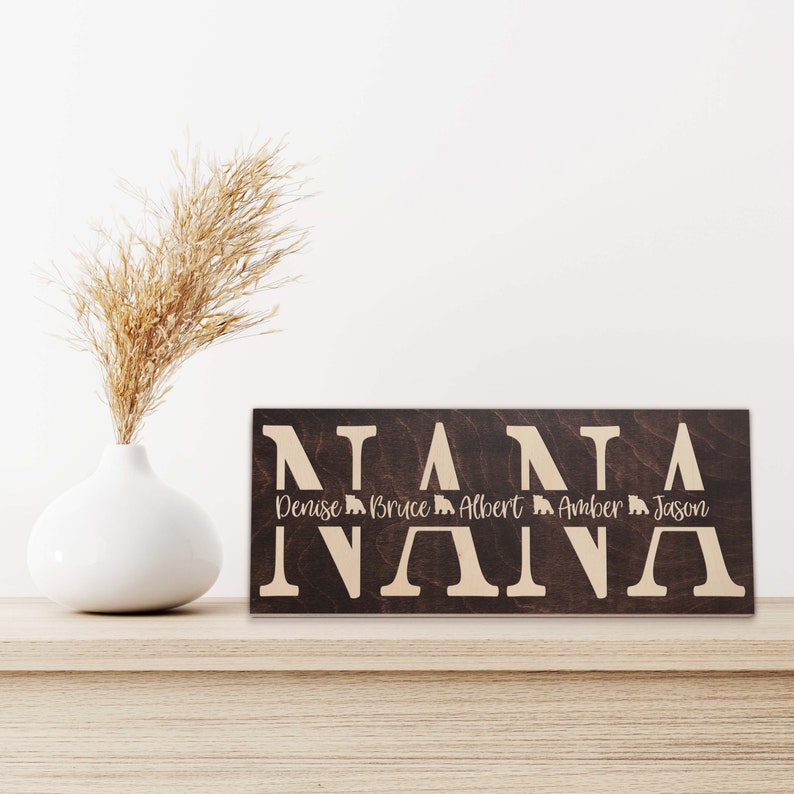 May include: A dark brown wooden sign with the word "NANA" in large white letters. Below the word "NANA" are the names "Denise, Bruce, Albert, Amber, Jason" in smaller white letters.