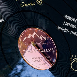 May include: A black vinyl record with a rose gold circle in the center. The circle has a mountain range design and the text "Our Adventure Begins" with the names "THE WILLIAMS" and "MARIAH CAREY" and the date "03.11.2024". The record also has the text "CONGRATS ON FINDING YOUR UNPAID THERAPIST!" on the right side.