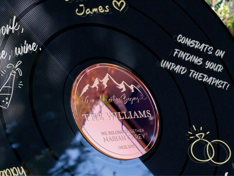 May include: A black vinyl record with a rose gold circle in the center. The circle has a mountain range design and the text "Our Adventure Begins" with the names "THE WILLIAMS" and "MARIAH CAREY" and the date "03.11.2024". The record also has the text "CONGRATS ON FINDING YOUR UNPAID THERAPIST!" on the right side.