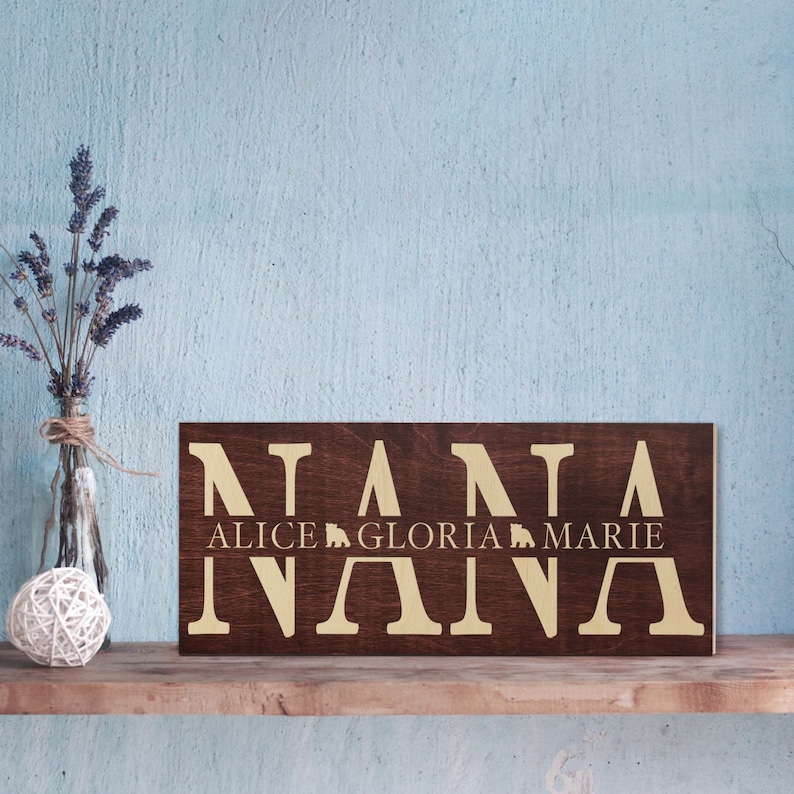May include: A dark brown wooden sign with the word "NANA" in large gold letters. Below the word "NANA" are the names "ALICE", "GLORIA", and "MARIE" in smaller gold letters.
