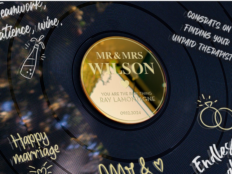May include: A gold-colored vinyl record with a gold mirror disc that says "MR & MRS WILSON" with a heart and the text "YOU ARE THE BEST THING RAY LAMONTAGNE 09.12.2024". The record has black and white handwritten text around the edge, including "Teamwork, Patience, Wine", "Happy Marriage", "Congrats on finding your unpaid therapist", and "Endless Love".