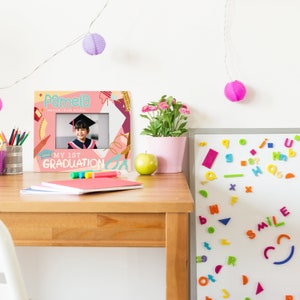 May include: A pink and colourful graduation photo frame with a photo of a young girl wearing a graduation cap and gown. The frame has the text "Pamela" and "My 1st Graduation" on it.