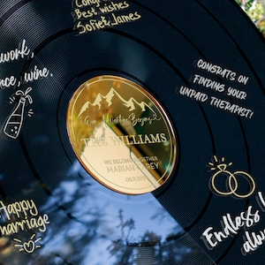 May include: A black vinyl record with gold lettering and a gold mirror insert. The record has the text "Teamwork, patience, wine." and "Happy marriage" written on it. The mirror insert has the text "Our Adventure Begins" and "The Williams" written on it. The record also has the text "Congrats on finding your unpaid therapist!" and "Endless love, always." written on it.