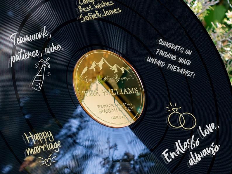 May include: A black vinyl record with gold lettering and a gold mirror insert. The record has the text "Teamwork, patience, wine." and "Happy marriage" written on it. The mirror insert has the text "Our Adventure Begins" and "The Williams" written on it. The record also has the text "Congrats on finding your unpaid therapist!" and "Endless love, always." written on it.