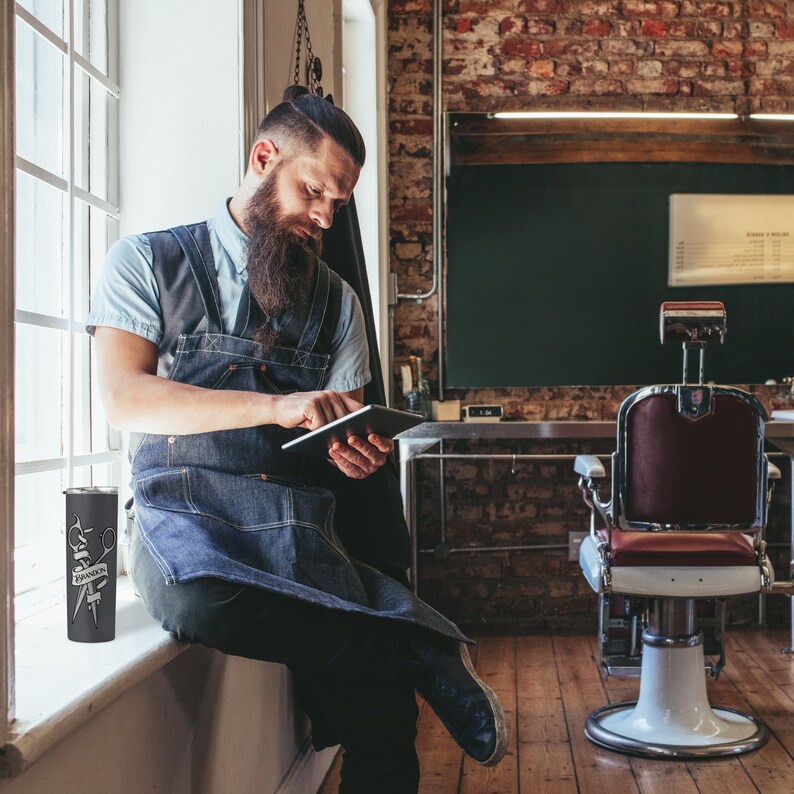 May include: A bearded man wearing a blue denim apron and a light blue shirt sits on a windowsill in a barbershop. He is holding a tablet and looking at the screen. A black and silver tumbler with a graphic of scissors and a razor is on the windowsill next to him.