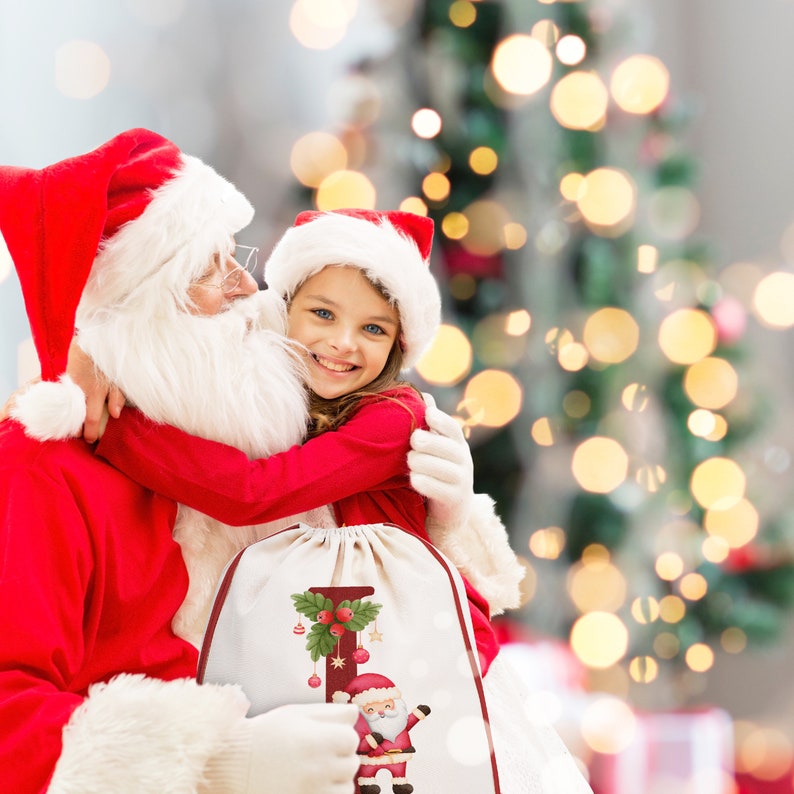 May include: Santa Claus in a red suit holds a young girl in a red dress. The girl is smiling and hugging Santa. They are standing in front of a Christmas tree with white lights. A white drawstring bag with a red and green Christmas design and the letter "I" is in front of them.