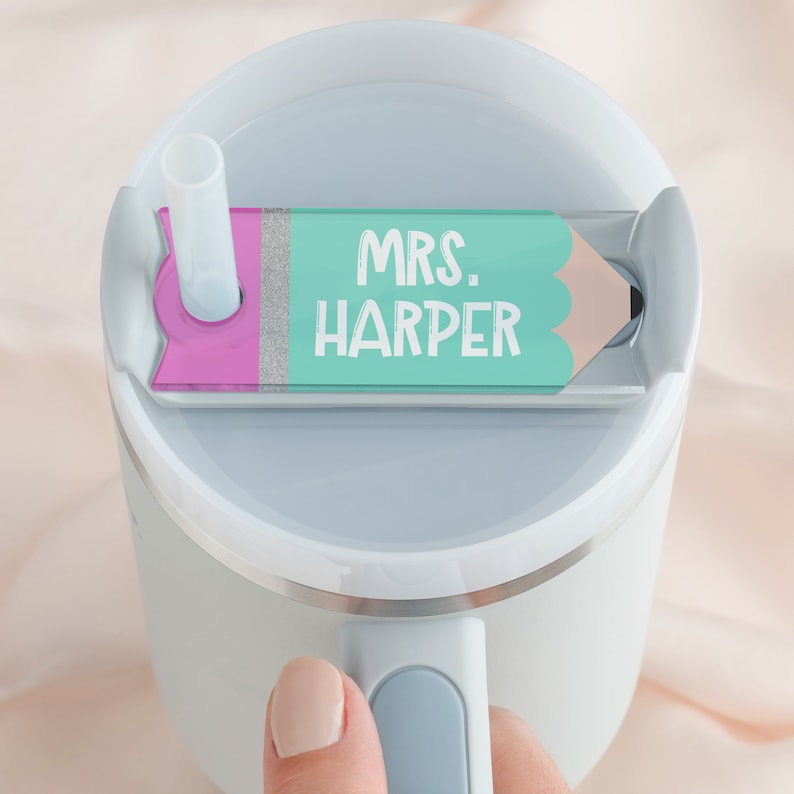May include: A white travel mug with a light blue lid and handle. The lid has a pink and green pencil-shaped tag that says "MRS. HARPER".