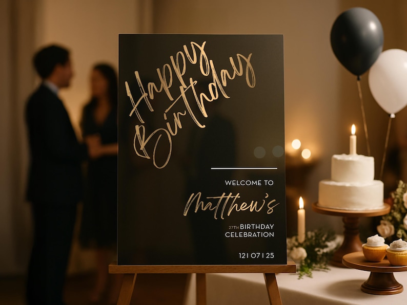 以下が含まれることがあります： 「Happy Birthday」と「Matthew's 27th Birthday Celebration」と金色の文字で書かれた黒い看板。背景には、ろうそくの灯った白いケーキと風船があります。