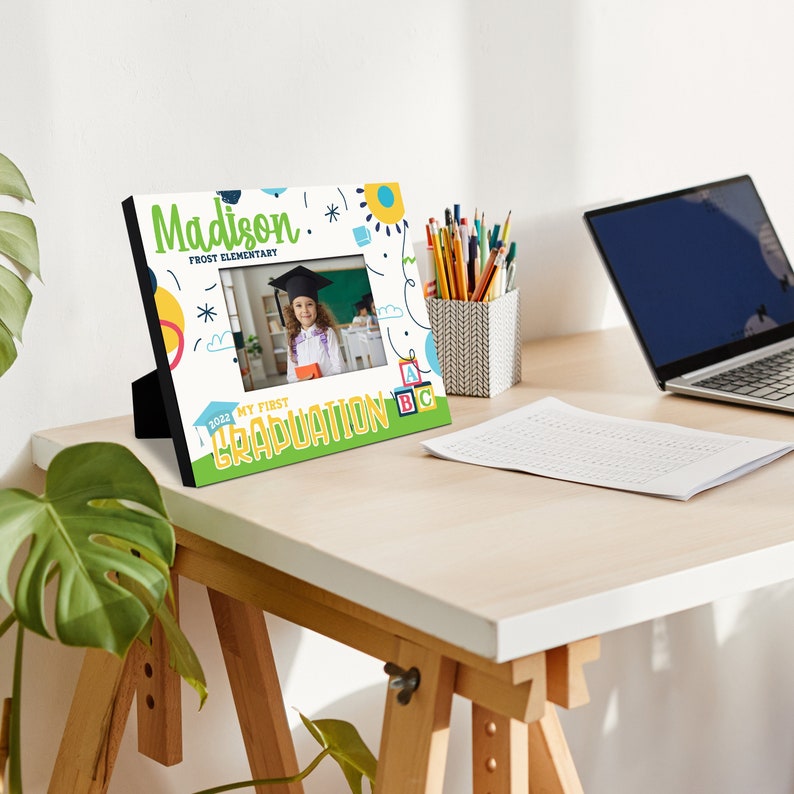 May include: A white picture frame with a colourful design featuring a child in a graduation cap and gown. The frame says "Madison Frost Elementary" and "My First Graduation 2022".