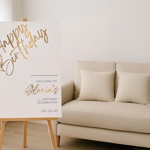以下が含まれることがあります： 「Happy Birthday」と「Gloria's 40th Birthday Celebration」と金色の文字で書かれた白い看板。看板は木製のイーゼルに立てられています。背景には、2つのクッションが付いたベージュのソファがあります。