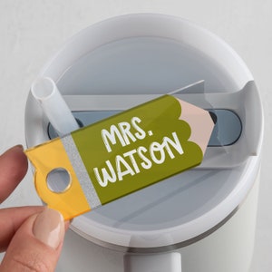 May include: A white travel mug with a green and yellow pencil-shaped tag that says "MRS. WATSON".