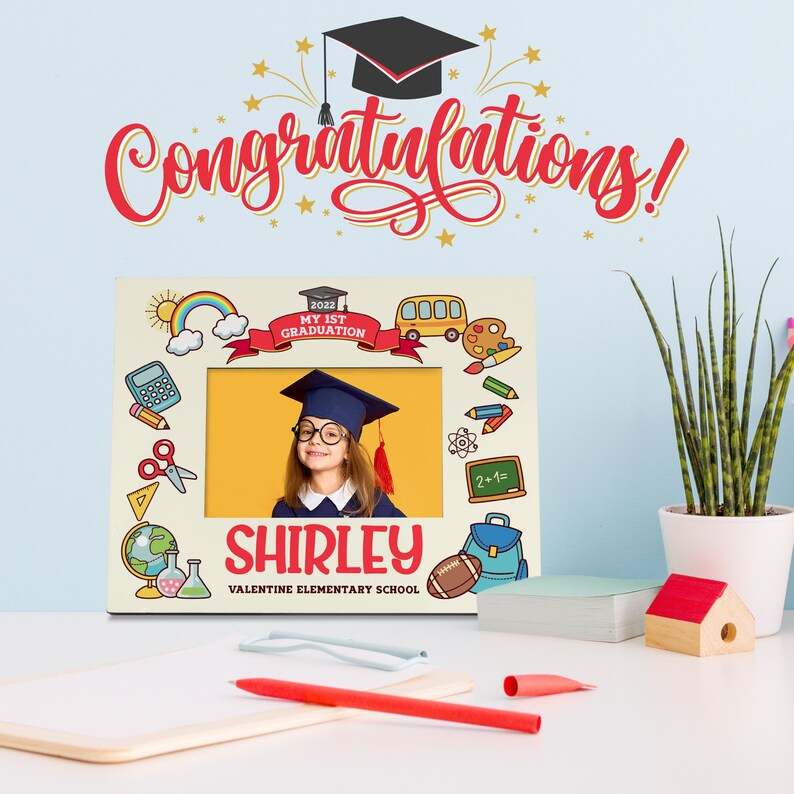 May include: A personalised graduation photo frame with a yellow background and colourful school-themed illustrations. The frame has a space for a photo and includes the text "2022 My 1st Graduation", "Shirley", and "Valentine Elementary School".