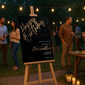 以下が含まれることがあります： 「Happy Birthday」と「Welcome to Michelle's 2nd Birthday Celebration」と金色の文字で書かれた黒い看板が木製のイーゼルに立てられています。看板は、人々やストリングライトのある屋外パーティーにあります。
