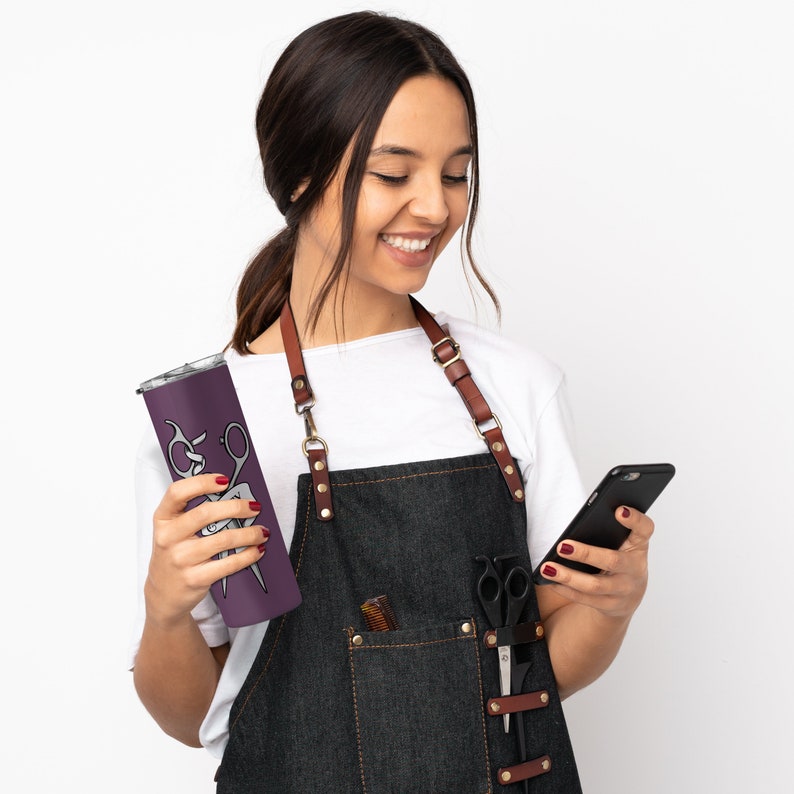 May include: A person wearing a white shirt and a dark blue denim apron with brown leather straps. The apron has a pocket with a pair of scissors and a comb. The person is holding a purple stainless steel tumbler with a graphic of a pair of scissors and the text "Hair Today, Gone Tomorrow". The person is also holding a black smartphone.