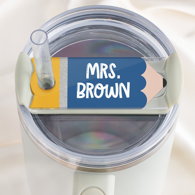 May include: A white travel mug with a blue and yellow pencil-shaped lid. The lid has a silver glitter stripe and the text "MRS. BROWN" on it.