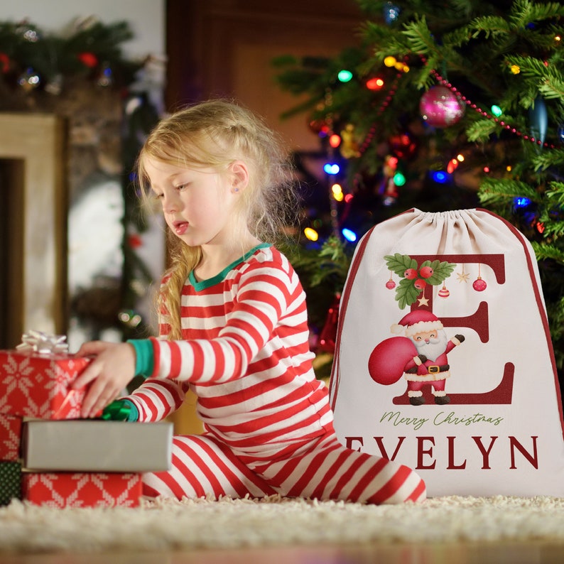 May include: A red and white personalized Christmas sack with the letter "E" and the name "Evelyn" printed on it. The sack features a cartoon Santa Claus holding a red bag and a green holly sprig with red berries. The text "Merry Christmas" is printed below the name.