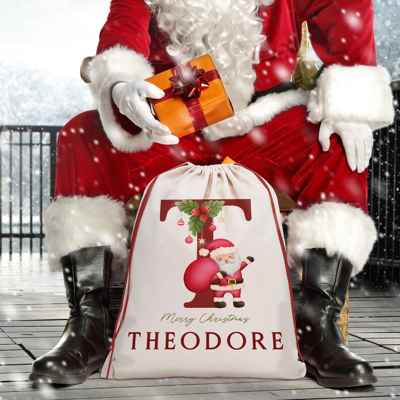 May include: A white canvas Santa sack with a red and green illustration of a Santa Claus holding a bag of gifts. The letter "T" is in the background with a green wreath and ornaments. The text "Merry Christmas THEODORE" is printed on the sack.