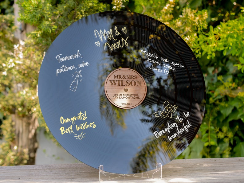 May include: A black vinyl record with a gold inscription that reads "Happily ever after starts today. James." and "Everyday to be wonderful." The record also has a gold inscription that reads "Teamwork, patience, wine." and "Congrats! Best wishes." A wooden plaque with the text "MR & MRS WILSON" is attached to the record. The plaque also has the text "YOU ARE THE BEST THING RAY LAMONTAGNE 0912.3004".