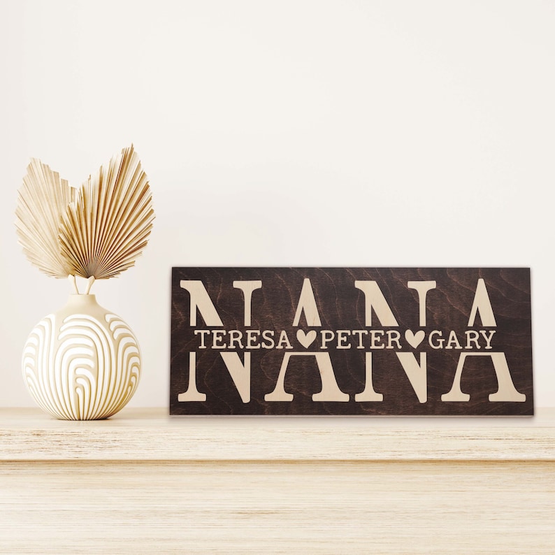 May include: A dark brown wooden sign with the word "NANA" in large white letters. Below the word "NANA" are the names "TERESA", "PETER", and "GARY" with a heart symbol between each name.