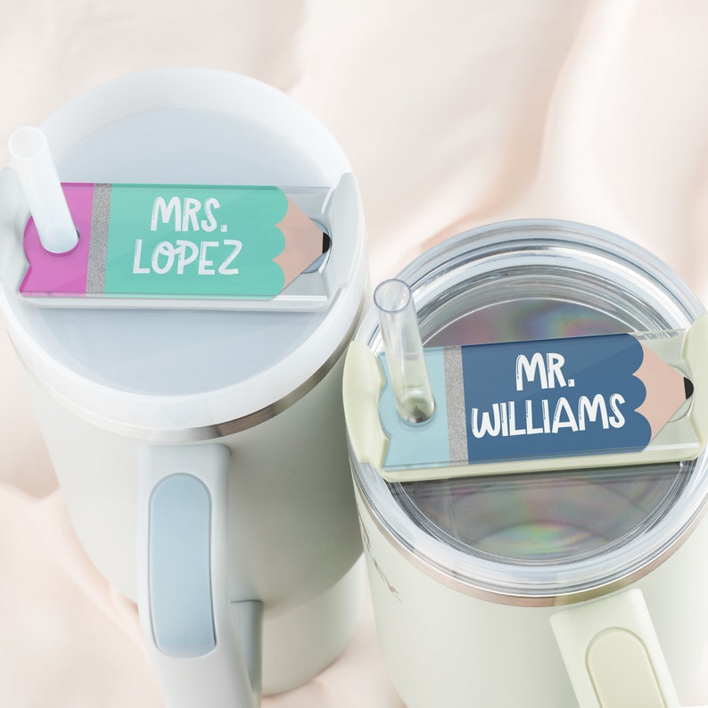 May include: Two white travel mugs with lids and straws. Each mug has a pencil-shaped name tag attached to the lid. The name tag on the left mug says "MRS. LOPEZ" in green and pink lettering. The name tag on the right mug says "MR. WILLIAMS" in blue lettering.