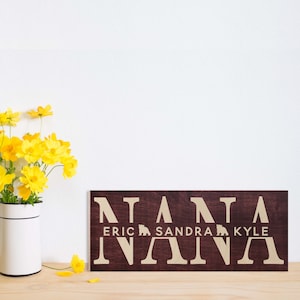 May include: A dark brown wooden sign with the word "NANA" spelled out in large white letters. Below the word "NANA" are the names "ERIC", "SANDRA", and "KYLE" in smaller white letters, with a small bear icon between each name.