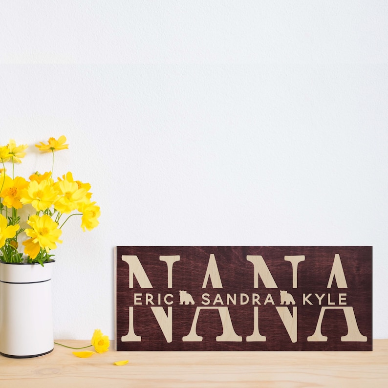 May include: A dark brown wooden sign with the word "NANA" spelled out in large white letters. Below the word "NANA" are the names "ERIC", "SANDRA", and "KYLE" in smaller white letters, with a small bear icon between each name.
