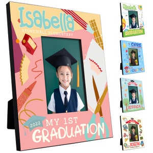 May include: A pink and yellow graduation photo frame with a colourful design featuring a graduation cap, scissors, and pencils. The frame has a black border and a cutout for a 4x6 inch photo. The text "My 1st Graduation" is printed on the frame.