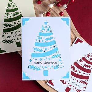 Peut inclure: Trois cartes de No&euml;l avec un motif d&eacute;coup&eacute; d'un sapin de No&euml;l. Les cartes sont blanches avec un motif d'arbre vert, bleu et rouge. Le texte sur les cartes est "Joyeux No&euml;l".