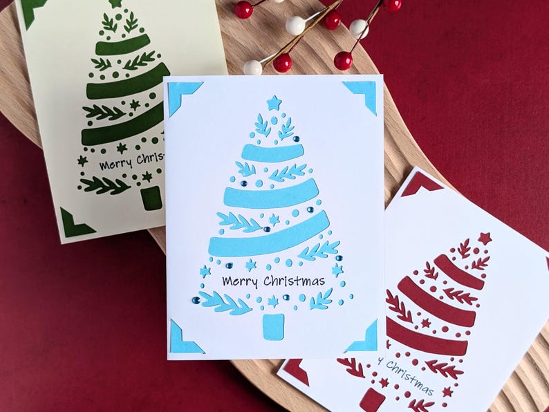 Peut inclure: Trois cartes de No&euml;l avec un motif d&eacute;coup&eacute; d'un sapin de No&euml;l. Les cartes sont blanches avec un motif d'arbre vert, bleu et rouge. Le texte sur les cartes est "Joyeux No&euml;l".