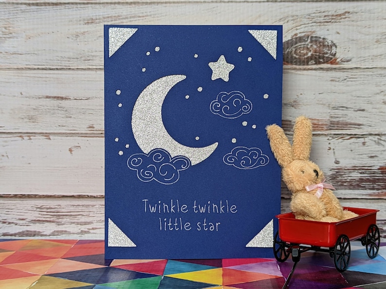 May include: A blue card with silver glitter accents features a crescent moon, a star, and clouds. The card has the text "Twinkle twinkle little star" printed on it. A small stuffed bunny sits in a red wagon.