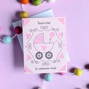 May include: A pink card with a white envelope. The card has a pink and black illustration of a baby carriage with a baby inside. The text on the card reads "Peek-a-boo to someone new!"