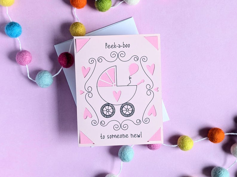 May include: A pink card with a white envelope. The card has a pink and black illustration of a baby carriage with a baby inside. The text on the card reads "Peek-a-boo to someone new!"