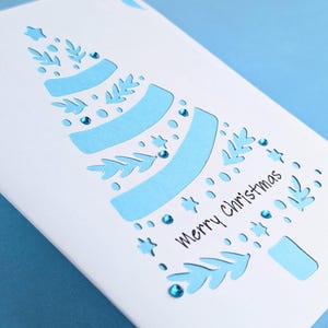 Peut inclure: Une carte blanche avec une d&eacute;coupe en papier bleu d'un sapin de No&euml;l. L'arbre est d&eacute;cor&eacute; de paillettes bleues et les mots "joyeux No&euml;l" sont &eacute;crits sur la carte.