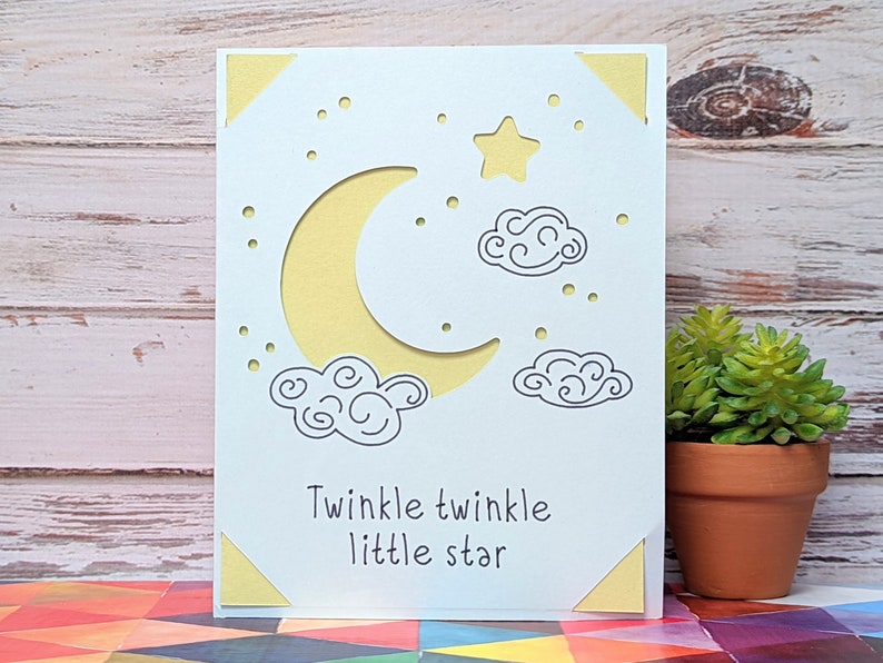 May include: A white card with a yellow crescent moon, a yellow star, and two gray clouds. The card has the text "Twinkle twinkle little star" written on it.