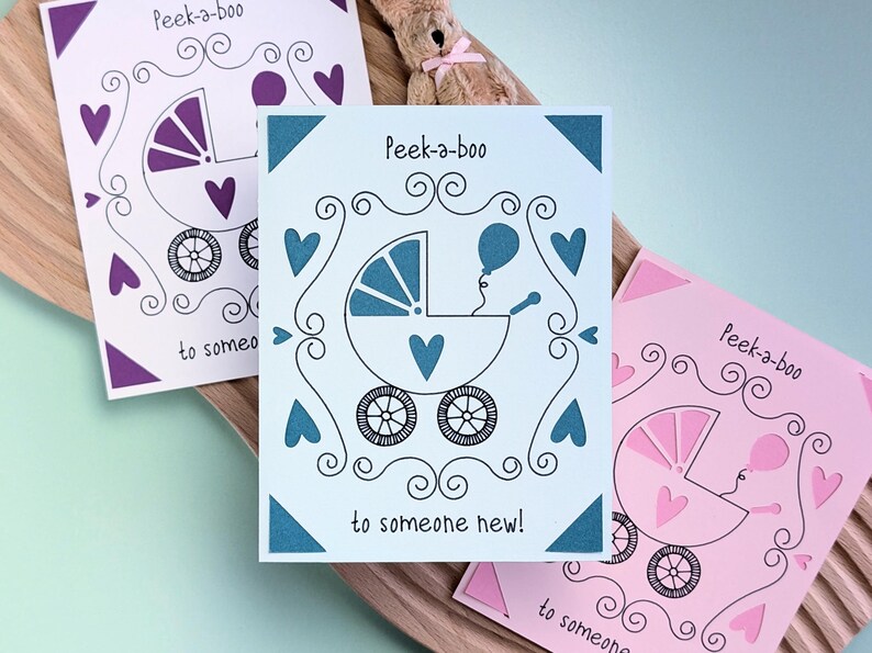 May include: Three baby shower cards with a peek-a-boo design. Each card features a stroller with a heart and a balloon. The strollers are colored purple, teal, and pink.