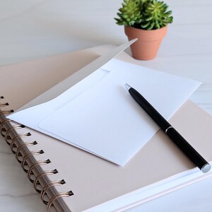 May include: A white envelope with a black pen on top of a spiral bound notebook with a light pink cover.