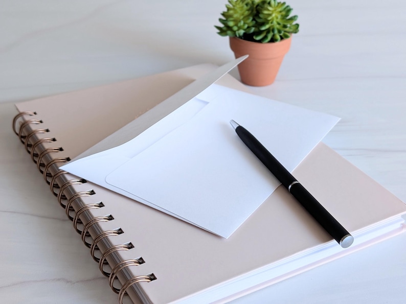 May include: A white envelope with a black pen on top of a spiral bound notebook with a light pink cover.