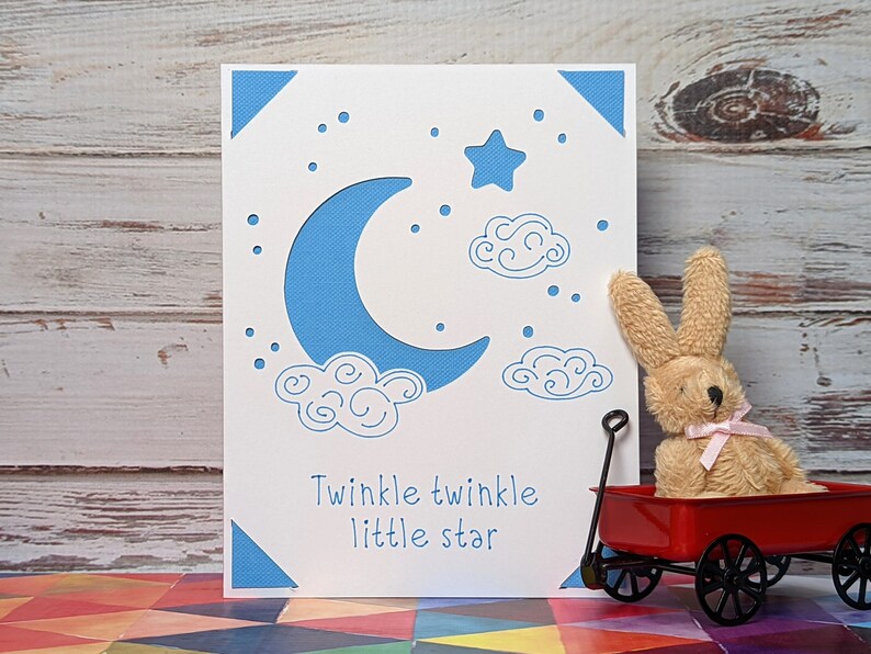 May include: A white card with a blue crescent moon, a white star, and white clouds. The card has the text "Twinkle twinkle little star" on it. A small red wagon with a brown bunny in it is in the foreground.