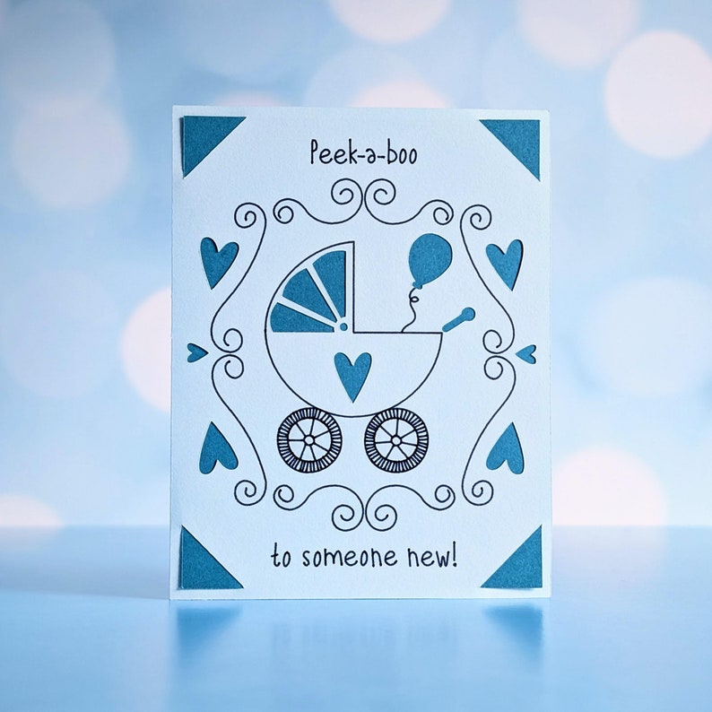 A cricut joy insert card with cutouts of a balloon, baby buggy, and hearts.  There are also ink drawings to complete the image.  The wording reads, "Peek-a-boo to someone new!"