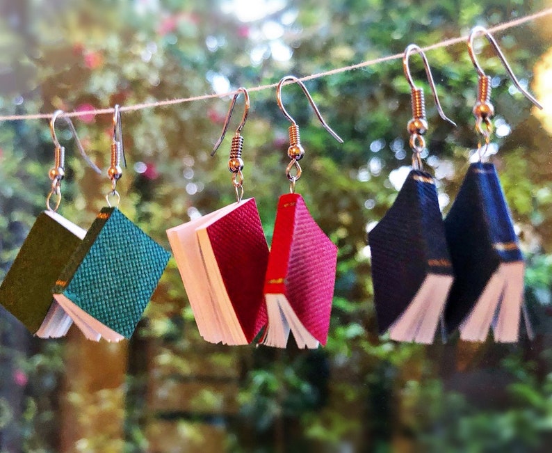 Op de afbeelding: Handgemaakte oorbellen in de vorm van miniatuurboeken, in verschillende kleuren zoals groen, blauwgroen, rood en marineblauw. De oorbellen hangen aan zilverkleurige haken en tonen de kleine boekontwerpen met gedetailleerde pagina's en omslagen.