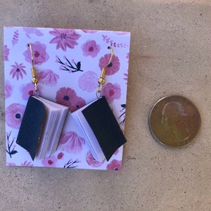 Op de afbeelding: Een paar handgemaakte oorbellen in de vorm van een boek met goudkleurige haken. De miniatuurboeken hebben donkerblauwe omslagen en witte pagina's. De oorbellen worden gepresenteerd op een kaart met bloemenpatroon.