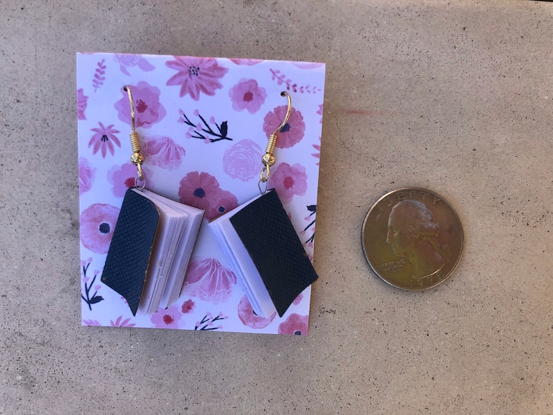 Op de afbeelding: Een paar handgemaakte oorbellen in de vorm van een boek met goudkleurige haken. De miniatuurboeken hebben donkerblauwe omslagen en witte pagina's. De oorbellen worden gepresenteerd op een kaart met bloemenpatroon.