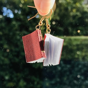 Op de afbeelding: Oorbellen in de vorm van een boek. Elke oorbel heeft een miniatuurboekje met een getextureerde, bordeauxrode kaft en witte pagina's. De boeken hangen aan goudkleurige oorhaken, wat een uniek literair accessoire cre&euml;ert.